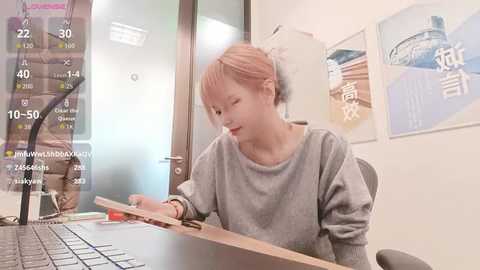 Video of an Asian woman with light skin and pink hair, wearing a gray sweater, sitting at a desk, using a laptop and a keyboard, in a brightly lit room with white walls and water bottles.