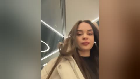 Video of a young woman with long brown hair, fair skin, and full lips, wearing a beige jacket, standing in a modern, dimly lit room with metallic walls and a glowing circular light.