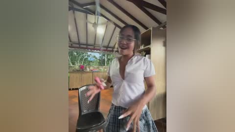 Video of a smiling, slender woman with glasses, wearing a white blouse and plaid skirt, standing in a rustic kitchen with wooden beams, a black chair, and a counter with pink flowers.