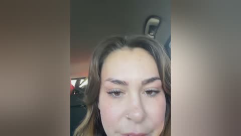 A close-up video of a woman with light skin, brown hair, and light makeup, showing her forehead and eyebrows. The background is blurred, featuring a car interior with a steering wheel.