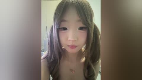 A close-up video of an East Asian woman with light skin, pigtails, and a delicate, youthful face. She's wearing a strapless top and has a small tattoo on her chest. The background is blurred, suggesting an indoor setting.