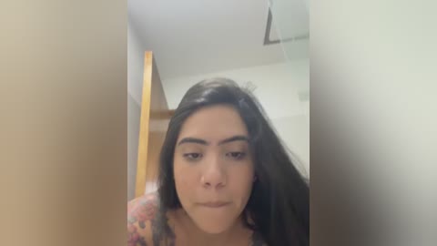 A close-up video of a young woman with long, dark hair and light skin, wearing a colorful top, standing in a dimly lit, narrow hallway with a wooden doorframe.