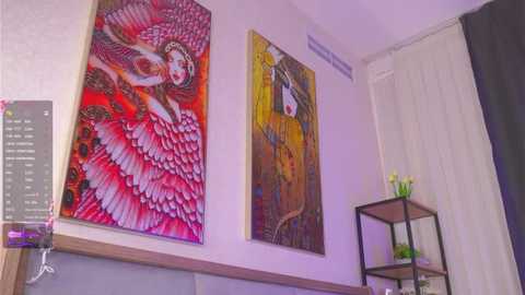 Video of a modern room featuring vibrant, surreal paintings of women with flowing red and pink feathered wings, hanging on a white wall. A black metal shelf with yellow flowers and a green vase is placed against a white curtain.