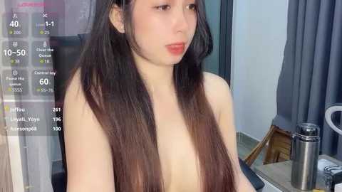 A video of a young Asian woman with long, straight brown hair, fair skin, and small breasts, sitting topless in a modern, dimly lit room.