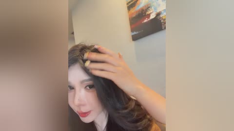A video of a woman with long, wavy black hair, fair skin, and red lipstick, touching her hair with a gold ring, in a modern, minimalist room with a colorful abstract painting on the wall.