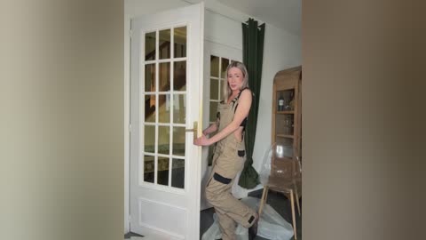 A video of a slender, blonde woman in tan overalls, standing at a white-framed glass door, in a dimly lit, beige-walled room with a wooden chair and a green curtain visible.