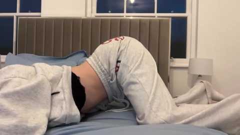 Video of a person in light gray sweatpants and a white sweatshirt, lying on a bed with blue sheets, facing away from the camera. The background features a beige upholstered headboard and a white bedside table with a lamp.