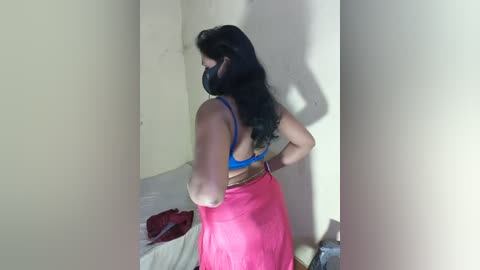 Media: Video of a woman with long black hair, wearing a blue bra and pink skirt, standing in a dimly lit room.