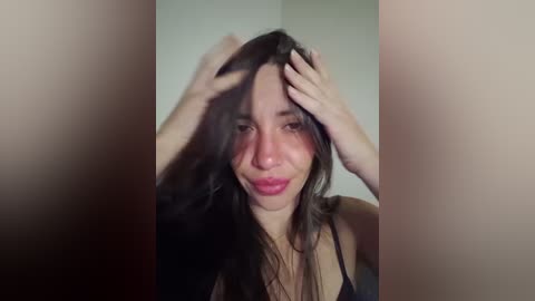 A close-up video of a young woman with long brown hair, fair skin, and a slight smile, wearing a black top, with her hands in her hair, taken indoors.