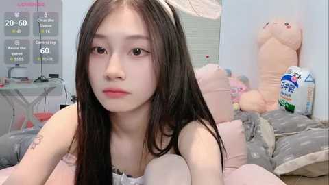 Video of an Asian woman with long black hair, wearing a white tank top, lying on a pink bed with stuffed animals and a bottle of shampoo in the background.