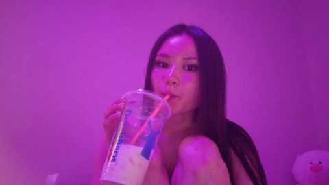Video of an Asian woman with long black hair, wearing a black top, drinking from a clear plastic cup with a pink straw, against a purple background.