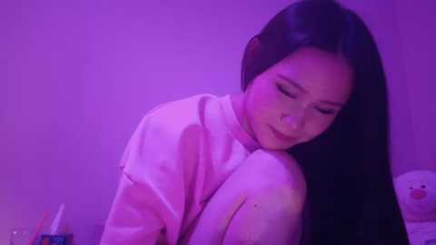 Video of an Asian woman with long black hair, wearing headphones, smiling, with a soft pink and purple lighting. She is in a cozy, possibly bedroom setting.