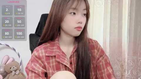 Media: Video of a young Asian woman with long, straight brown hair, wearing a red plaid shirt, sitting in a chair, looking thoughtful. Background includes a calendar and stuffed toys.