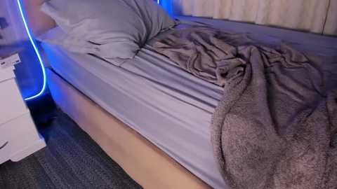 Video of a neatly made bed with a light grey fitted sheet, a grey pillow, and a brown, fuzzy blanket. The room has blue LED strip lighting, a white nightstand, and a beige curtain in the background.