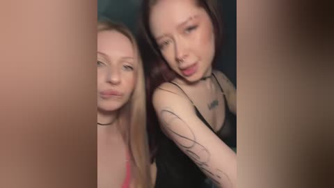 Video of two young women with fair skin, one with long blonde hair and the other with long dark hair. They are wearing black tank tops, with tattoos visible on their arms. Background is blurry, making them the focal point.