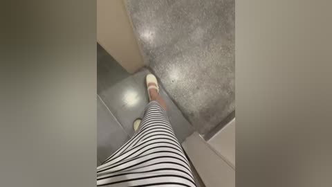 A video from a low angle shows a person's legs and feet in a striped dress, standing on a carpeted floor with beige walls in the background.