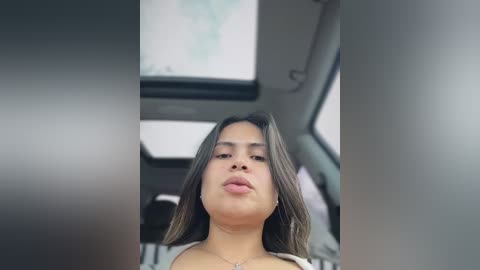 Video of a young Latina woman with medium brown skin, long brown hair, and a confident expression. She's wearing a light-colored top. The background shows an airplane cabin with a blurred map screen.