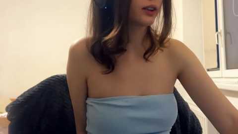 A video of a young woman with fair skin, wearing a strapless light blue top, sitting on a dark textured couch. She has long, wavy brown hair, and is talking into a headset, with a window in the background.