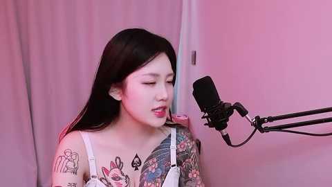 Video of a young East Asian woman with long black hair, wearing a white top, singing into a microphone. She has colorful tattoos on her shoulders and chest. Background is a pink wall and curtain.