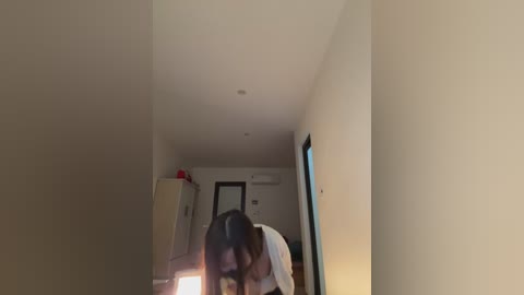 A video of a narrow, dimly-lit hallway with beige walls. A person with dark hair and a white shirt leans forward, casting a shadow. A white fridge and a red item are on the left, with a doorway and air conditioning unit visible in the background.