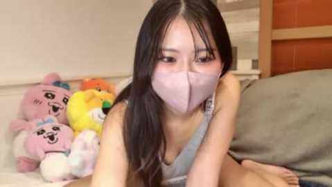 A video of an Asian woman with long black hair, wearing a light blue face mask, lying on a bed, surrounded by colorful stuffed animals.