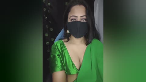 Video of a South Asian woman with medium skin tone, wearing a black face mask, green satin blouse, and blue hijab. Background features dark curtains with green lights.