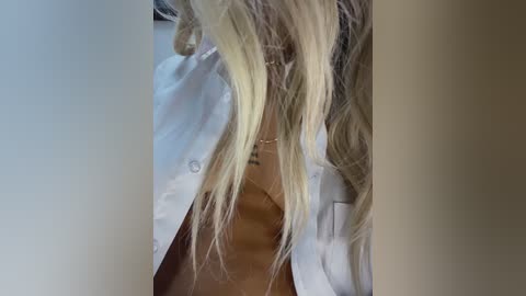 Video of a blonde woman with wavy hair, wearing a white button-up shirt, partially unbuttoned, revealing cleavage, against a blurred background.