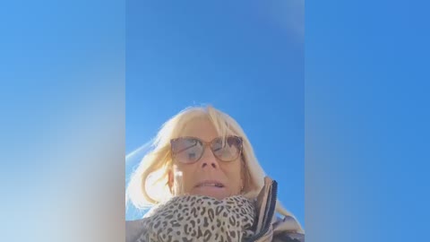 Video of a blonde woman with large sunglasses, looking upward into a clear blue sky. She wears a leopard print scarf, with a background that emphasizes the bright sky.