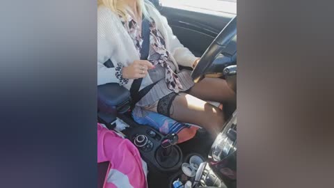 Video of a woman in a car, wearing a white sweater and patterned skirt, driving with a seatbelt on, holding a phone, with a pink jacket on the passenger seat.