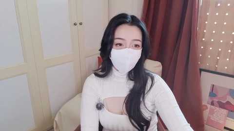 Video of an Asian woman with long black hair and fair skin wearing a white mask and turtleneck sweater, standing in a cozy room with beige curtains and a soft light background.