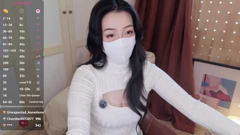 Video of an Asian woman with long black hair, wearing a white face mask and tight white top, sitting on a beige sofa. Background shows a framed artwork and a window with curtains.