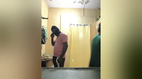 Video of a large, dark-skinned man with a beard, wearing a pink t-shirt and black shorts, taking a mirror selfie in a small, cluttered bathroom with a yellow shower curtain.