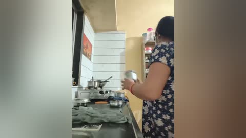 Video of a kitchen with a woman in a floral dress stirring a pot on a stove. The background features white tiled walls and a beige cabinet, with utensils scattered on the countertop.