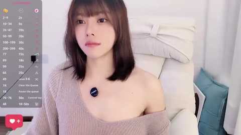 Video of an East Asian woman with shoulder-length brown hair, wearing a beige off-shoulder knit sweater, sitting on a white chair in a room with white walls.