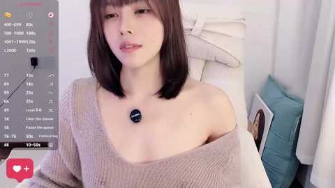 A video of a young East Asian woman with straight, shoulder-length brown hair and light skin, wearing a beige off-shoulder sweater. She has a small pendant necklace and sits on a white cushioned chair in a softly lit room.