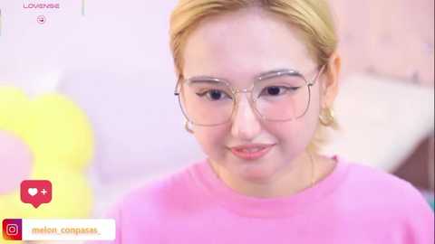 Video of a fair-skinned, blonde-haired young woman with glasses, wearing a pink shirt, smiling. Background features a pastel-colored room with a yellow heart-shaped object.