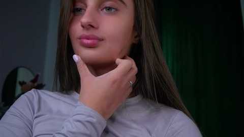 Video of a young woman with light brown skin and straight, shoulder-length brown hair, wearing a light gray long-sleeve top. She has a contemplative expression, with her hand gently touching her chin, and a green curtain in the background.