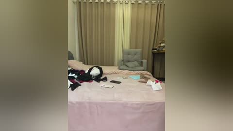 A video of a messy bedroom with beige curtains, a gray chair, a bed with a pink duvet, scattered clothes, and a smartphone on the floor.