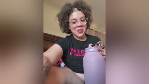 Video of a light-skinned woman with curly hair, wearing glasses and a black t-shirt with \"Don't Be A Bitch\" in pink, holding a purple water bottle, sitting indoors.