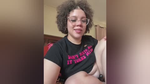 Video of a Black woman with a medium build, wearing a black crop top with \"I don't like my hair\" in pink letters, and glasses. She is sitting on a bed with a red blanket, smiling.