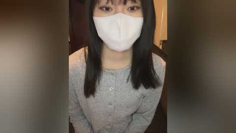 Video of an East Asian woman with long black hair, wearing a white face mask, gray cardigan, and standing indoors against a blurred background.