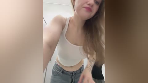 A video of a young woman with fair skin, long blonde hair, wearing a white crop top and grey jeans, smiling in a dimly lit room, partially obscured by a wooden frame.