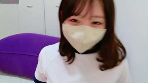 Video of an Asian woman with long dark hair, wearing a white lab coat and beige face mask, sitting on a purple exam table in a clinical setting.