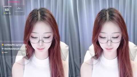 A video of a young Asian woman with long, straight, reddish-brown hair, wearing glasses and a white top, standing against a gray curtain background.