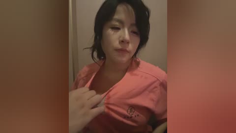Video of an Asian woman with short black hair, wearing a pink shirt, partially unbuttoned, in a dimly lit, beige-walled bathroom.