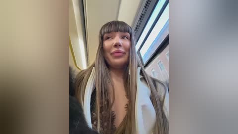 Video of a woman with long, straight brown hair and bangs, wearing a black dress, standing in a subway car with fluorescent lighting.