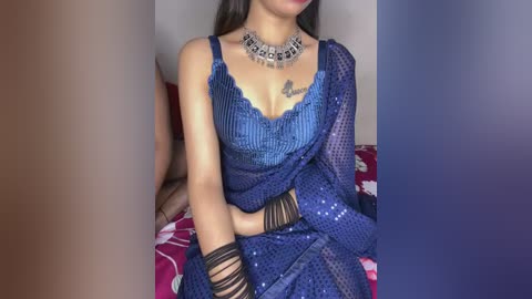 Video of a South Asian woman in a blue sequined saree and black bangles, seated on a bed with red and white floral sheets. She wears a silver choker necklace.