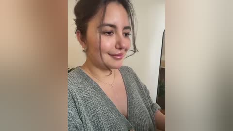 Video of a young Latina woman with light brown skin, dark hair in a messy bun, wearing a gray knitted V-neck sweater, smiling slightly, standing indoors with neutral-colored walls.