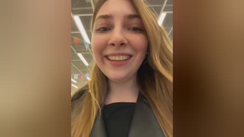 Video of a smiling young woman with long blonde hair, wearing a black top and a black jacket, standing in a brightly lit room with visible ceiling tiles and colorful banners in the background.