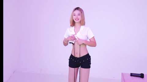 Video of a cheerful Asian woman with shoulder-length blonde hair, wearing a white crop top and black shorts, holding a microphone, against a white backdrop.